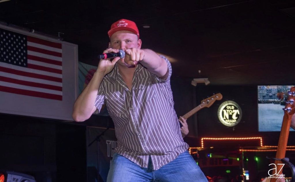 Male singer performing at Batesville Music Bash, part of Batesville Main Street events, with American flag in background and vibrant stage lighting.